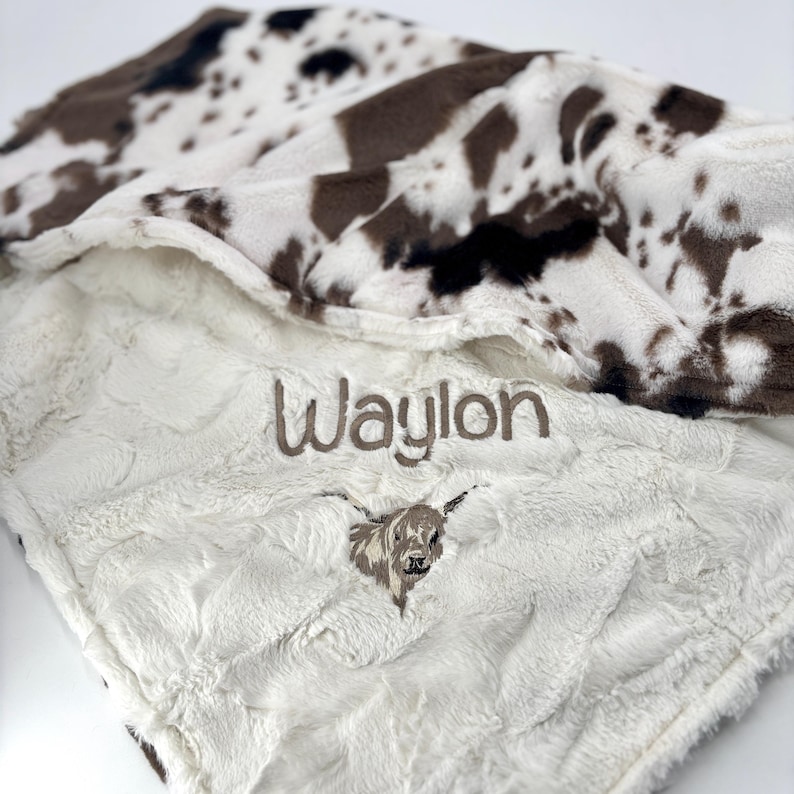 May include: A soft, white and brown cow print baby blanket. The blanket has the name "Waylon" embroidered in brown thread, along with an embroidered Highland cow. The blanket is made of a plush, textured material.