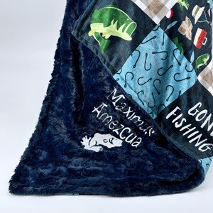 Puede incluir: Una manta azul marino y estampada con una superficie afelpada y texturizada. La manta presenta un diseño de patchwork con gráficos temáticos de pesca, incluyendo peces, anzuelos y el texto "Gone Fishing". También tiene texto bordado que dice "Maximus Amezcua".
