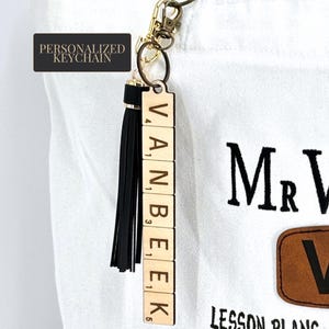 Może przedstawiać: Spersonalizowany brelok do kluczy z imieniem "VANBEEK" zapisanym drewnianymi kafelkami z literami. Brelok ma czarny frędzel i złote okucia. Tekst "PERSONALIZED KEYCHAIN" znajduje się w czarnym pudełku.