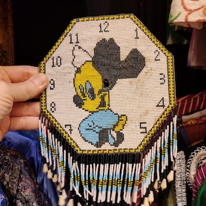 May include: A white octagonal beaded clock featuring a cartoon duck with a black hat and yellow beak. The clock face has black numbers 1 to 12. The clock has a fringe of black, white and blue beads.