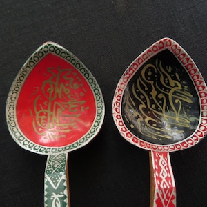 Puede incluir: Dos cucharas de madera con diseños intrincados. Una cuchara es roja con caligrafía dorada, la otra es negra con caligrafía dorada. Ambas cucharas tienen bordes verdes y blancos.
