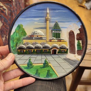 May include: A round, hand-painted decorative plate featuring a detailed relief of a Middle Eastern architectural scene. The plate has a black border and depicts a mosque with a minaret, domes, and a green lawn. The color palette includes shades of yellow, green, and blue.