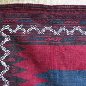 May include: Close-up of a red, black, and blue woven textile with a white geometric pattern. The textile has a fringed edge.