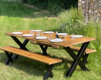 May include: A wooden picnic table with two benches, set up outdoors on a grassy lawn. The table has a black X-shaped base and features built-in ice coolers. The table is set with white plates, glasses, and a bottle of wine.