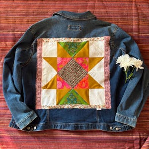 May include: A blue denim jacket with a colorful patchwork design on the back. The patchwork features a star pattern with pink, green, gold, and white fabric. Pink lace trim borders the patchwork.