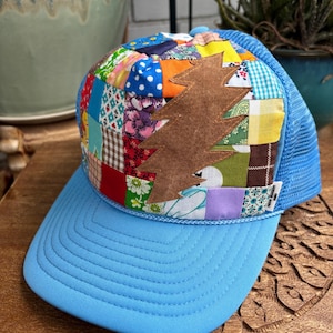 May include: A trucker hat featuring a patchwork front panel made of various colorful fabric squares in floral, gingham, and polka dot patterns. A brown suede lightning bolt applique is centered on the front. The brim and mesh back are light blue.