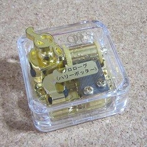May include: A gold-colored music box with a clear plastic case. The music box has a winding key shaped like a treble clef. The label on the music box reads "Melody (Music Box)".