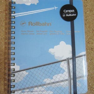 May include: A blue-covered spiral-bound notebook with a sky design, including clouds, an aeroplane, and a fence. The "Rollbahn" logo is displayed, alongside "Campus" and "Rollbahn" on a black tag. A black elastic band secures the notebook.