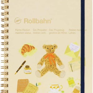 以下が含まれることがあります： クリーム色のRollbahnノート。金色のスパイラル綴じとネイビーブルーのゴムバンド留め。表紙には、テディベア、クロワッサン、ペン、インクボトル、本、ティーカップ、糸巻きのイラストが描かれています。「Rollbahn」の文字が見えます。