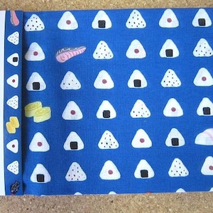 Op de afbeelding: Blauwe stof met een herhalend patroon van witte driehoeken met verschillende ontwerpen. De stof heeft Japanse tekst die "おにぎり" leest, wat zich vertaalt naar "onigiri" of "rijstbal".