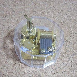 May include: A gold-colored music box with a clear plastic lid. The music box is in the shape of an octagon and has a winding key on the top.
