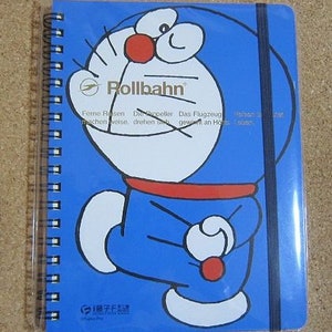 May include: A blue spiral bound notebook with a cartoon character on the cover. The character is blue with a red mouth and a white outline. The text "Rollbahn" is printed in white on the cover. The text "Forne Rosen Die Propeller Das Flugzeug" is printed in white on the cover.