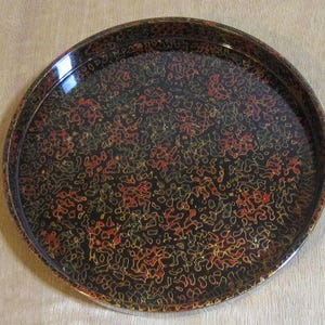 May include: A circular black tray with a glossy finish. The tray features a pattern of red, yellow, and green organic shapes. It has a raised edge and a reflective surface, suitable for serving.