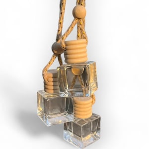 May include: Three glass car air fresheners hanging from a natural twine string with wooden beads. The fresheners are square and clear, with a light brown liquid inside.