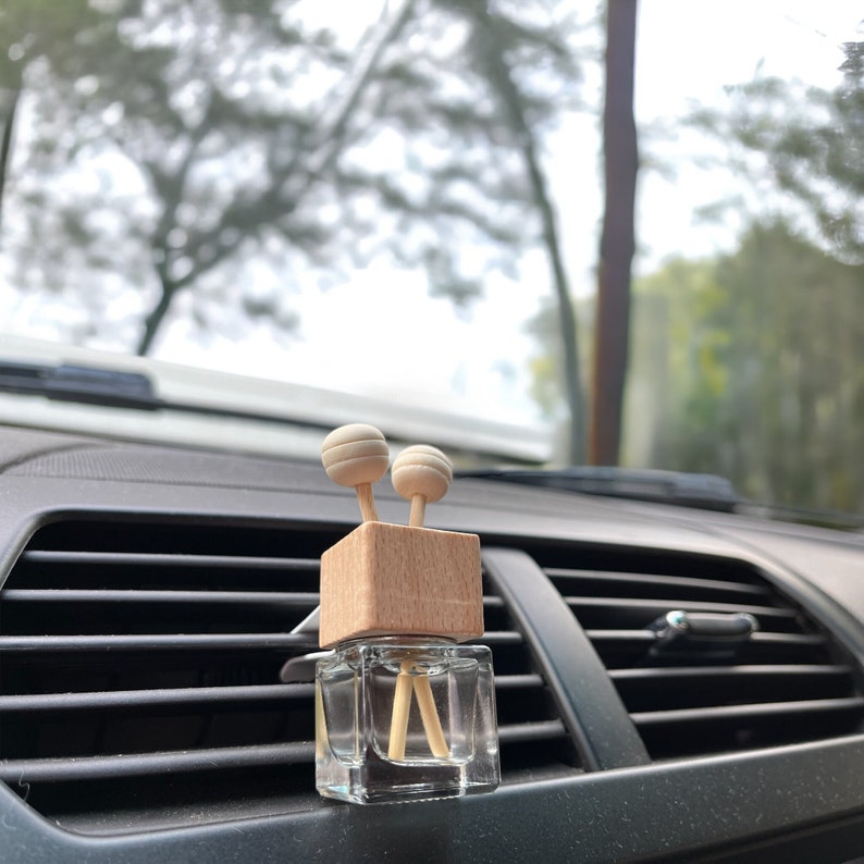 A wooden car vent diffuser clipped onto a car air vent. The clear glass bottle contains fragrance oil, with wooden reeds absorbing and dispersing the scent.