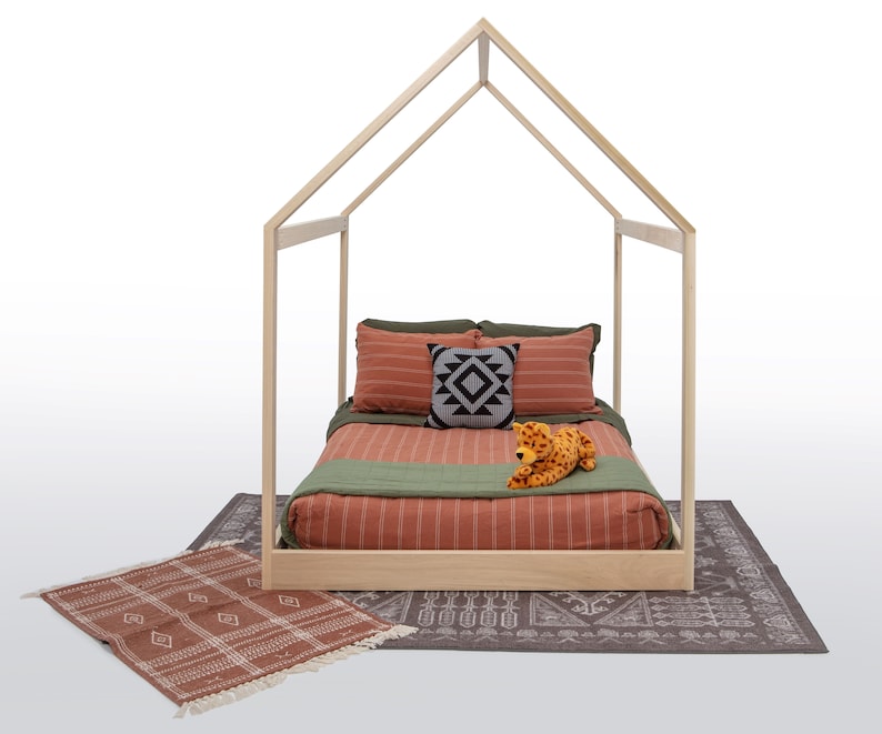 May include: A light-colored wooden house-shaped bed frame featuring a mattress with rust-colored striped bedding, olive green accents, a small square patterned throw pillow, and a plush leopard toy, placed on a dark patterned rug.