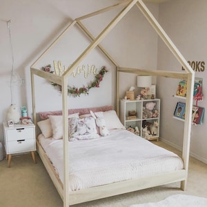 May include: A light-colored wooden house-shaped bed frame with a peaked roof structure. The bed is dressed with a light pink quilt, several pillows, and a plush rabbit toy. A white two-drawer nightstand sits to the left, and a white four-cube shelf unit is positioned to the right.