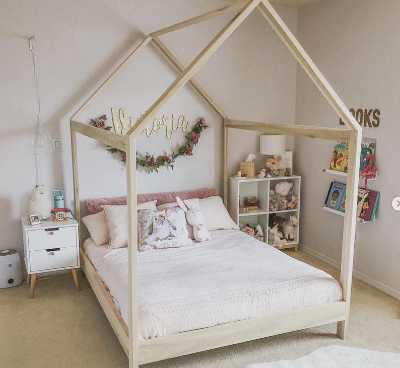 May include: A light-colored wooden house-shaped bed frame with a peaked roof structure. The bed is dressed with a light pink quilt, several pillows, and a plush rabbit toy. A white two-drawer nightstand sits to the left, and a white four-cube shelf unit is positioned to the right.
