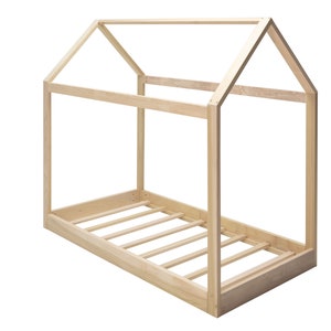 May include: A light-colored wooden bed frame shaped like a house. The structure features a rectangular base with horizontal slats and a peaked roof frame constructed from four vertical posts connected by beams at the top and sides.