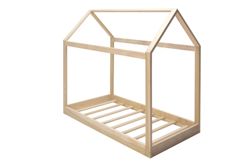 May include: A light-colored wooden bed frame shaped like a house. The structure features a rectangular base with horizontal slats and a peaked roof frame constructed from four vertical posts connected by beams at the top and sides.