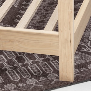 May include: A light-colored natural wood bed frame featuring a sturdy corner post and horizontal side rails. The structure includes a series of evenly spaced wooden slats designed to support a mattress, showcasing the grain of the unfinished wood.