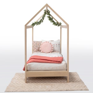 May include: A light-colored wooden house-shaped bed frame featuring a peaked roof structure draped with green artificial foliage. The bed is dressed with a white quilt, a coral-colored throw blanket, and several pillows, including a round pink pom-pom cushion.