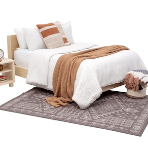 May include: A light wood bed frame with a white duvet and brown blanket. The bed has a brown and white patterned throw pillow and a tan throw pillow with orange accents. A small light wood nightstand with two shelves is next to the bed. The nightstand has a clock and a stack of books on it. A gray and white patterned rug is on the floor.
