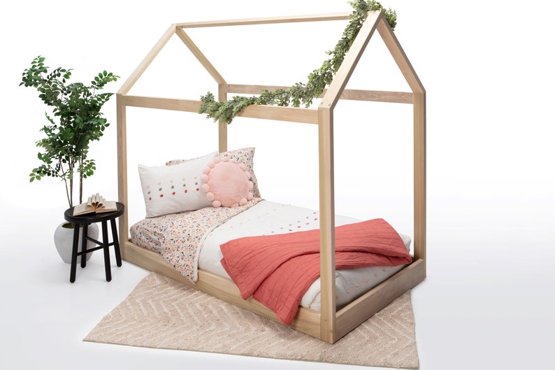 May include: A light-colored wooden house-shaped bed frame featuring a peaked roof design. The bed is dressed with white bedding, a floral patterned sheet, a round pink pom-pom pillow, and a solid coral-colored textured throw blanket.
