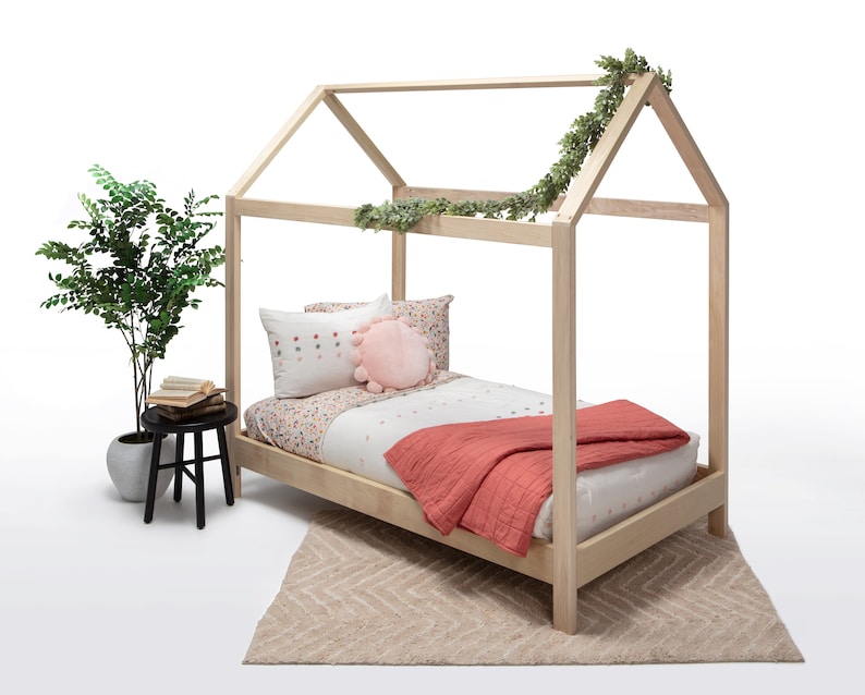 May include: A light-colored wooden house-shaped bed frame featuring a white mattress with a floral patterned sheet, a solid coral-colored quilt, a white pillow with small dots, and a round pink pom-pom pillow, decorated with a green vine.