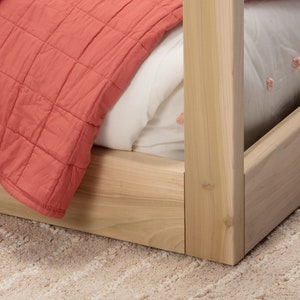 May include: A light-colored wooden bed frame corner featuring a square post and side rails. The bed is dressed with a white sheet featuring small pink embroidered details and a coral-colored quilted blanket draped over the side.