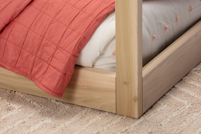 May include: A light-colored wooden bed frame corner featuring a square post and side rails. The bed is dressed with a white sheet featuring small pink embroidered details and a coral-colored quilted blanket draped over the side.