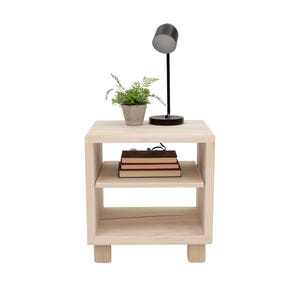 May include: A light-colored wooden nightstand with two shelves. A small potted plant, a black desk lamp, and a stack of books are on top. The nightstand has a simple, modern design.