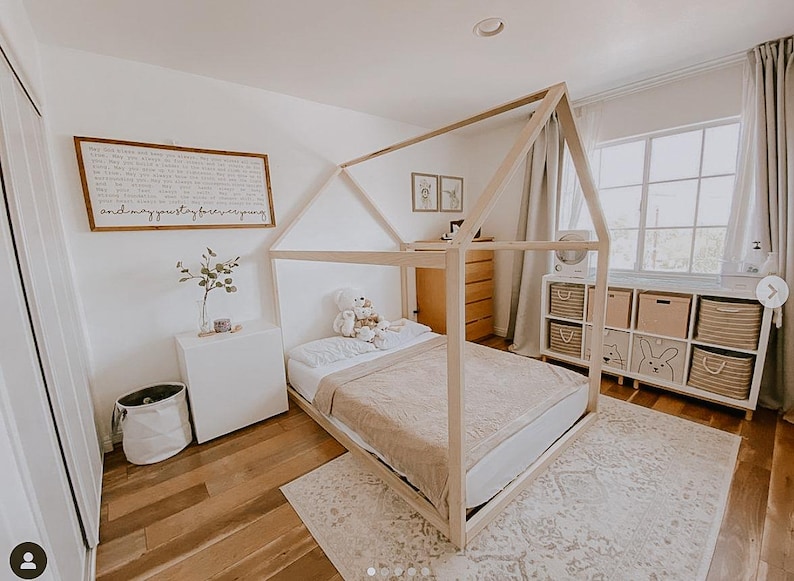 May include: A light-colored wooden house-shaped bed frame with a white mattress and a beige blanket. A white teddy bear sits on the bed. A white sign with black text hangs on the wall above a white side table with a small plant.