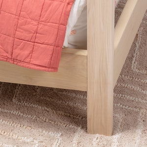 May include: A light-colored wooden bed frame corner featuring a square post and side rail. A coral-colored quilted blanket with a grid pattern is draped over the side, partially covering a white sheet tucked into the bed frame structure.