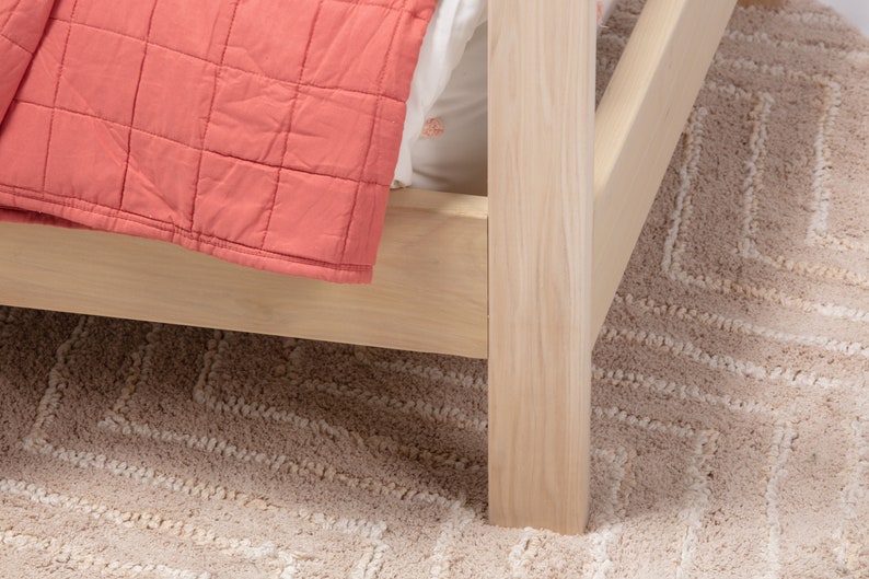 May include: A light-colored wooden bed frame corner featuring a square post and side rail. A coral-colored quilted blanket with a grid pattern is draped over the side, partially covering a white sheet tucked into the bed frame structure.