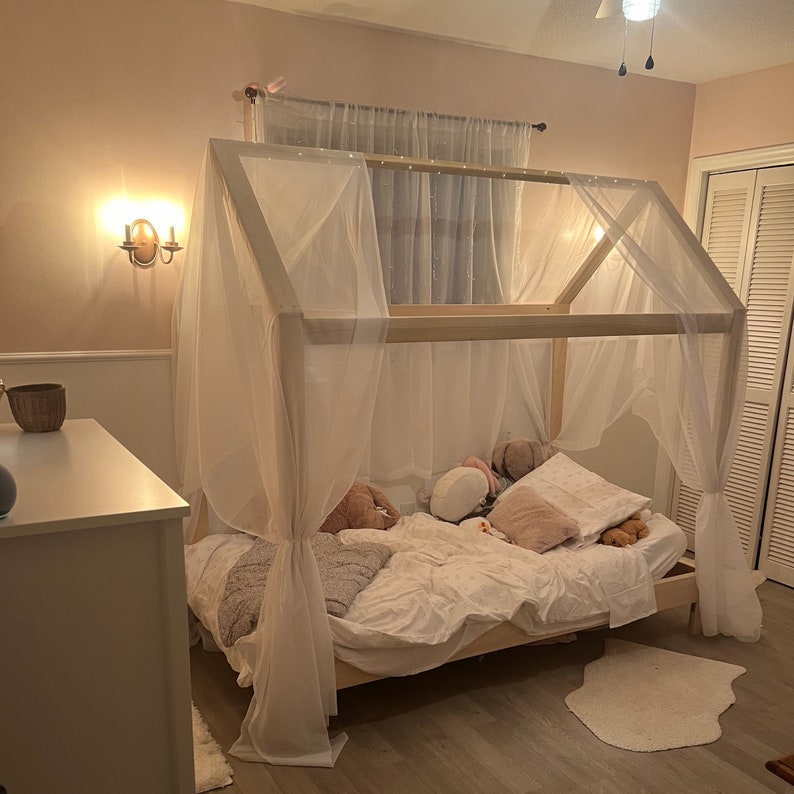May include: A light-colored wooden house-shaped bed frame featuring a peaked roof structure draped with sheer white fabric curtains. The bed is dressed with white bedding and several plush pillows, creating a soft and layered appearance.