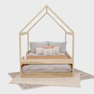 May include: A light-colored wooden house-shaped bed frame featuring a peaked roof structure. The bed is dressed with a gray duvet, a tan blanket, and several decorative pillows in shades of gray, white, and a small coral-colored square cushion.