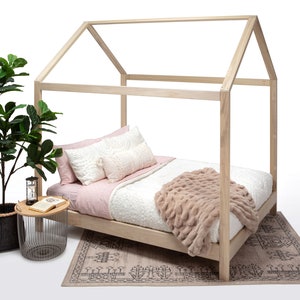 May include: A light-colored wooden house-shaped bed frame with a mattress, light pink and white pillows, and a textured beige throw blanket. Beside the bed is a small round metal side table with a book and an hourglass.