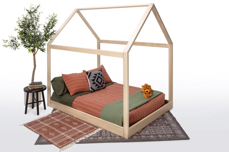May include: A light-colored wooden house-shaped bed frame featuring a mattress with rust-colored striped bedding, olive green sheets, and a small plush leopard toy. The bed is placed on a dark patterned rug, with a small black side table nearby.