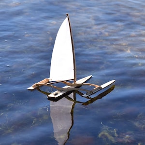 May include: A small wooden sailboat with a white sail floats on the water. The sailboat has a single mast and a hull with two outriggers.