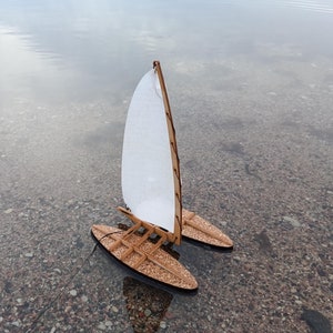 Könnte beinhalten: Ein kleines Holzsegelboot mit einem weißen Segel, das in seichtem Wasser schwimmt. Das Segelboot hat zwei Rümpfe und ist aus hellem Holz gefertigt.