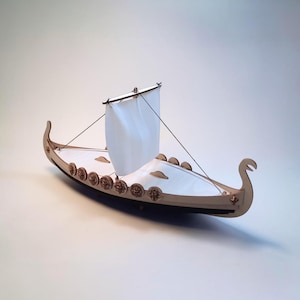 May include: A wooden model of a Viking ship featuring a light-colored hull with carved circular shield details along the sides, a white fabric sail attached to a mast, and a curved prow and stern. The structure is assembled from laser-cut wood.