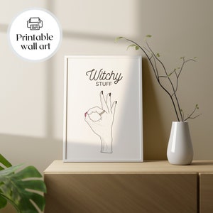 May include: Printable wall art with a white frame and a white background. The art features a black and white illustration of a hand making the OK sign with the text "Witchy Stuff" above it.