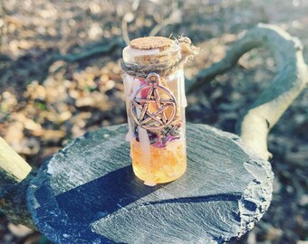 May include: A small glass bottle filled with crystals, herbs, and a pentagram charm. The bottle is sitting on a dark grey slate surface.