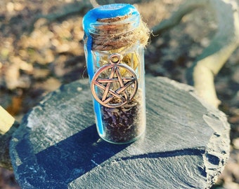 May include: A small glass bottle with a blue top and a cork stopper. The bottle is filled with dried herbs and a silver pentagram charm hangs from the cork.