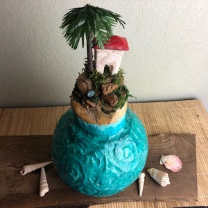 May include: A teal blue ceramic vase with a textured surface, depicting a miniature island with a small white house with a red roof, a palm tree, and moss. The vase is sitting on a wooden surface with seashells.