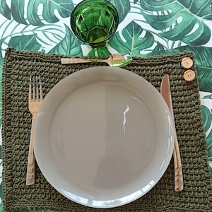 Peut inclure: Mise en place avec un set de table vert crocheté, une assiette beige et des couverts en bois. Un verre vert et deux boutons en bois sont également présents. Le fond présente un motif de feuilles tropicales.
