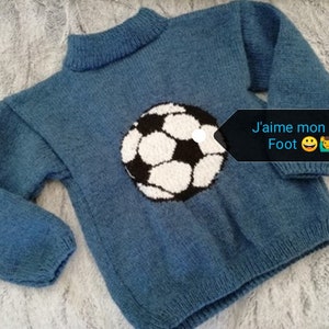 Peut inclure: Un pull bleu en tricot avec un motif de ballon de football blanc et noir. Le pull a un col roulé et des manches longues. Le texte "J'aime mon pull Foot" est écrit sur une étiquette blanche attachée au pull.