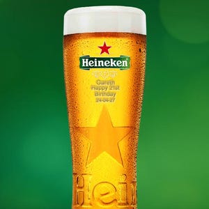 May include: A tall glass of Heineken beer, with condensation and a white foamy head. The glass has a star design and the Heineken logo. Text on the glass reads: "Gareth Happy 21st Birthday 24-04-27". The background is a solid green.