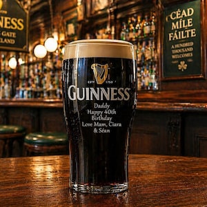 Op de afbeelding: Een pint Guinness stout met een romige schuimkraag, op een houten tafel in een pub. Het Guinness-logo en tekst zijn zichtbaar op het glas, samen met een verjaardagsbericht. De achtergrond toont een bar met flessen en een bord.